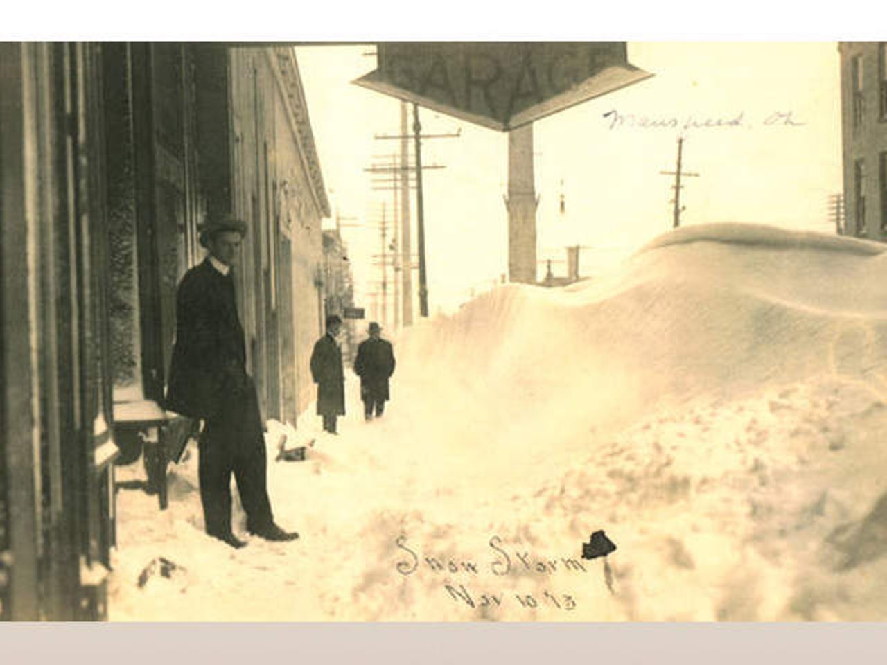 A man stands next to a large snowdrift, under a garage sign, after the Great Lakes Hurricane of 1913.