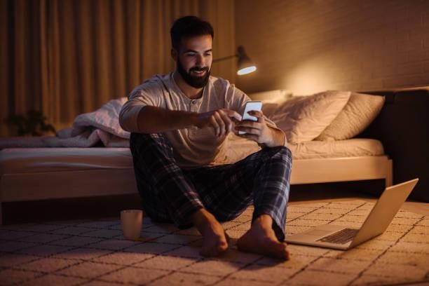 a man sitting beside his bed with a phone in his hand and a laptop in front of him